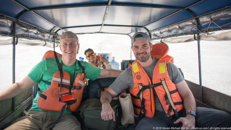 Two smiling passengers in orange life jackets seated inside a covered river launch, with another passenger behind them.