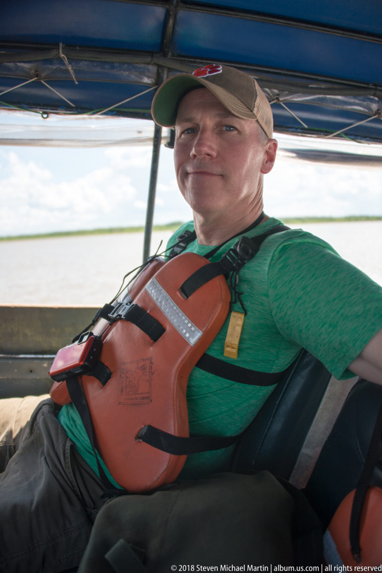 Seated passenger in green shirt and orange life jacket beneath the blue canopy of a small river launch.