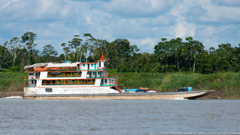 Multi-deck white, green, and orange river vessel underway on broad brown water with forested bank behind.