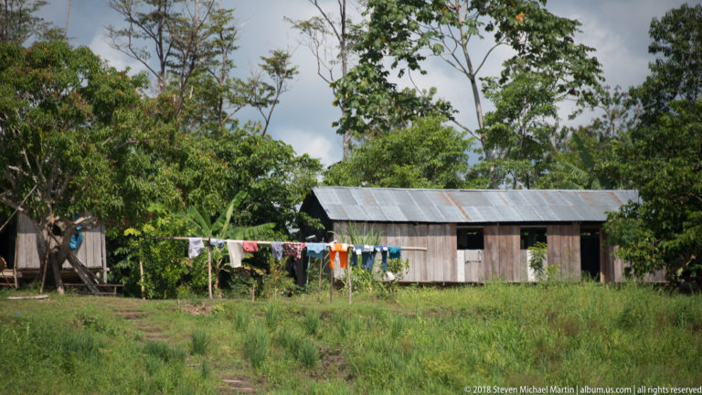 Long wooden riverside house with corrugated roof and laundry line in front of dense tropical vegetation.