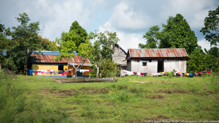 Wooden riverside houses with corrugated roofs, laundry lines, grass, and a narrow canoe in an Amazonian settlement.