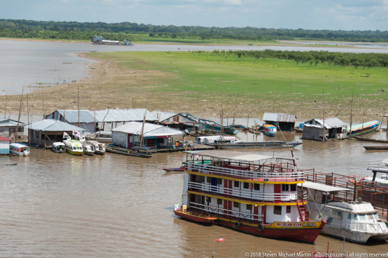 Large red, white, and yellow multi-deck riverboat beside low sheds and small boats on a shallow waterfront.