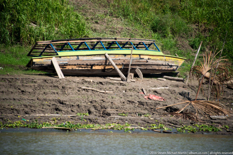 Long narrow wooden riverboat resting on a muddy riverbank beside tropical vegetation and a simple timber frame.