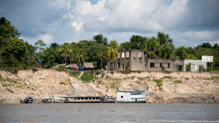 Small boat below a steep eroded river bluff topped by palms and simple masonry buildings near Iquitos.