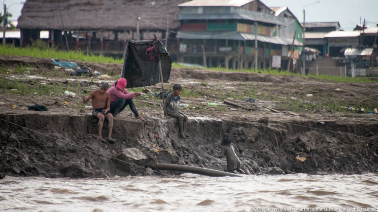 Several people beside a steep eroded muddy bank with a projecting pipe and riverside buildings behind.