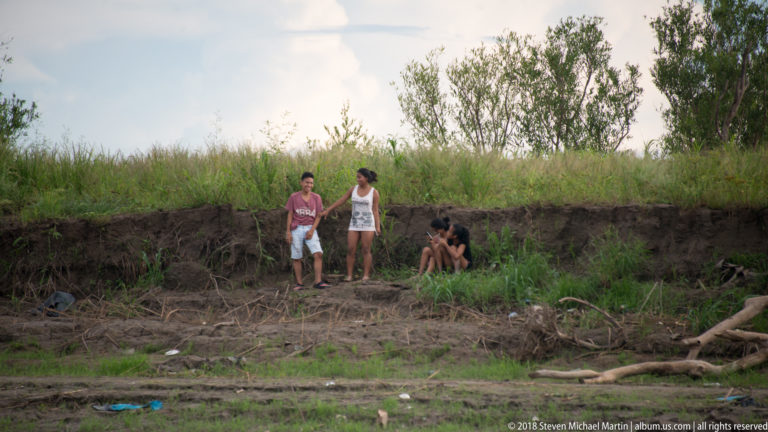 Small group standing above an eroded grassy riverbank with exposed soil and driftwood near Iquitos.