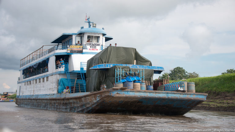 Blue-and-white river vessel Eduardo VIII carrying covered cargo and containers near the Iquitos riverbank.