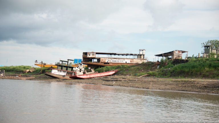 Several small riverboats hauled onto an exposed muddy bank beside simple waterfront workshops.
