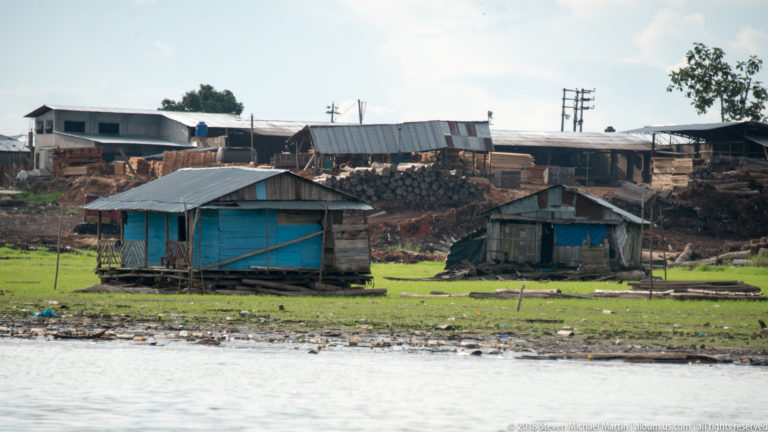 Small wooden stilt buildings beside stacked timber and sheds on the riverbank near Iquitos.