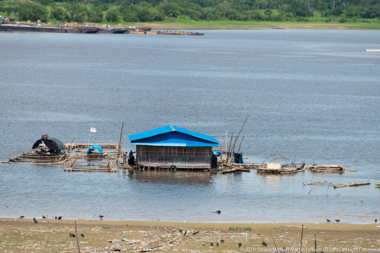 Blue-roofed floating or stilted riverside structure surrounded by narrow platforms, poles, and shallow water.