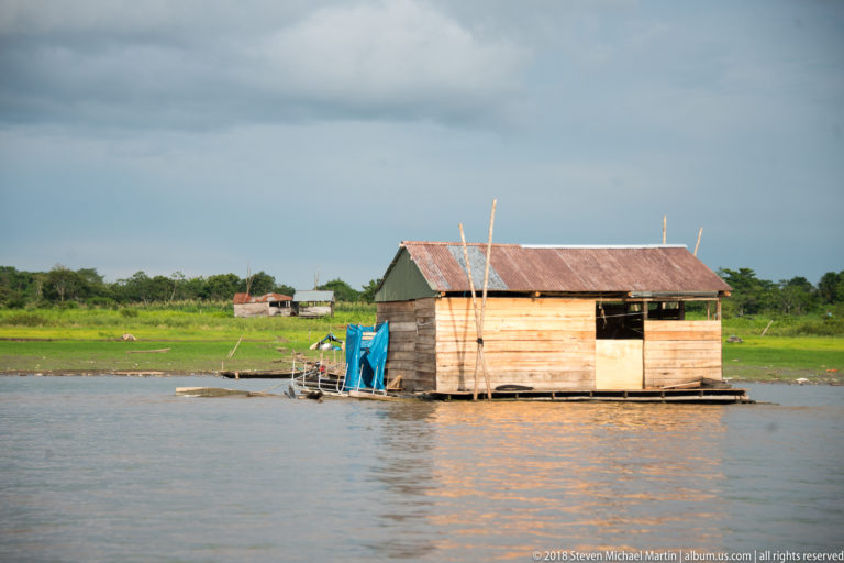 Simple timber house with corrugated roof standing on a low floating platform in the river.