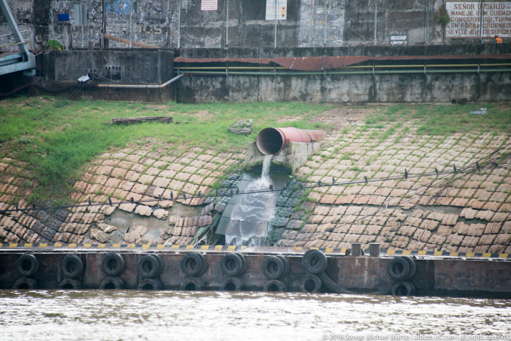 Large drainage pipe discharging water down a paved riverbank embankment beside the Iquitos waterfront.