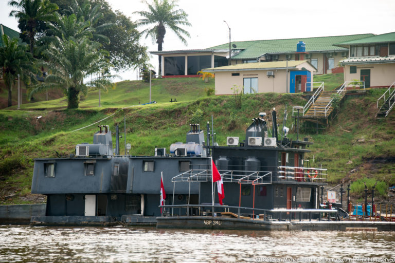 Dark utility boat flying a Peruvian flag moored below a developed riverbank facility.