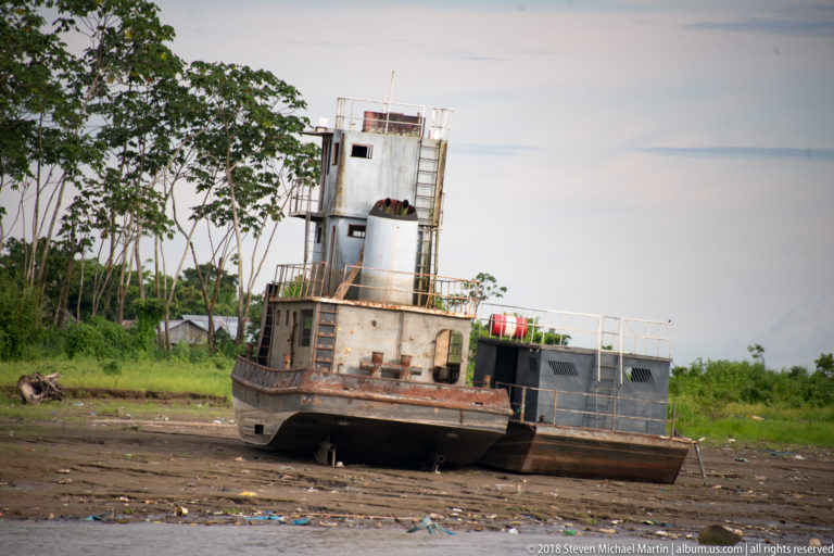 Small steel utility boat hauled onto the muddy riverbank with wheelhouse, ladders, and railings visible.