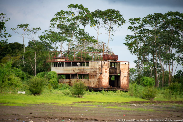Large rusting river-vessel hulk resting on a grassy bank beside dense tropical vegetation.