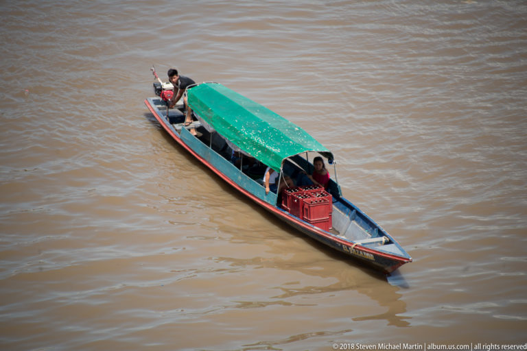 Long narrow motor canoe with green canopy carrying several people across brown river water.