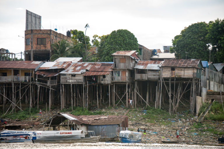 Timber riverside houses raised on tall posts above a muddy shoreline with small boats below.