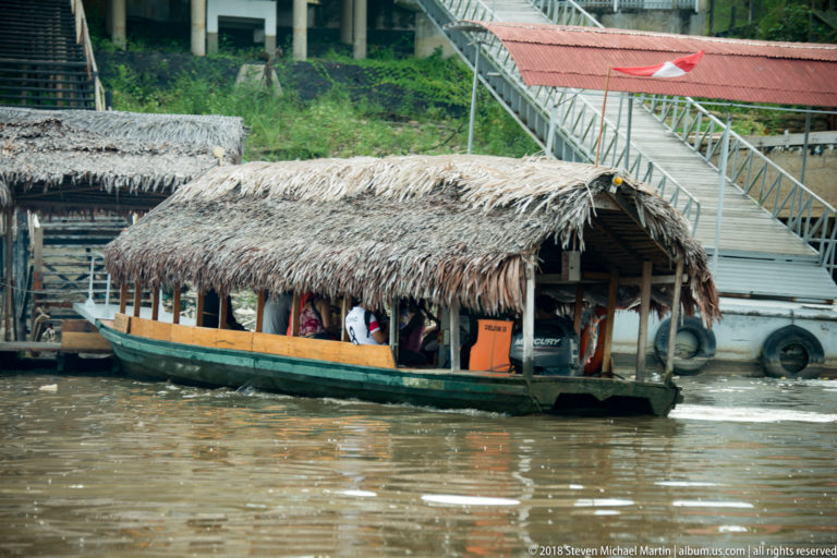Open-sided passenger boat with a palm-thatch roof moving beside the Iquitos docks.