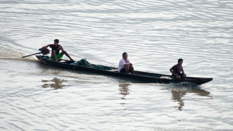 Three people ride in a long narrow canoe, one operating a small outboard motor at the stern.