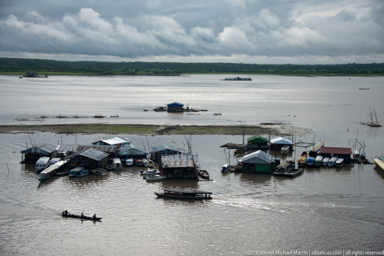 Narrow canoe with several people crossing in front of moored riverboats and floating structures.