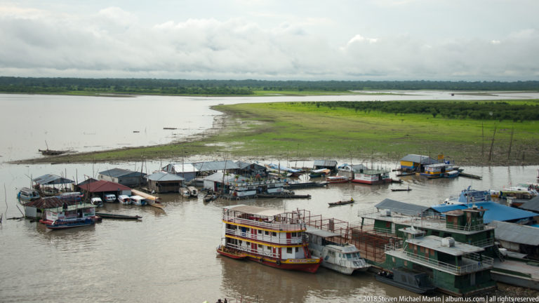 Clustered riverboats and dock structures below a broad green floodplain and cloudy sky near Iquitos.