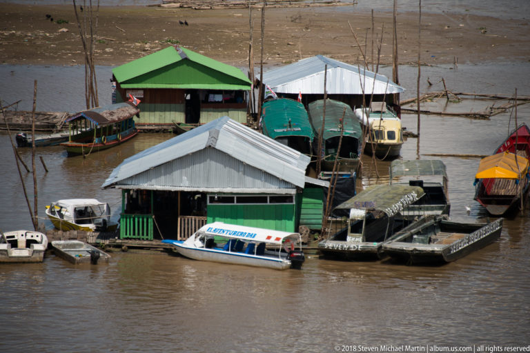 Cluster of floating waterfront buildings with green and metal roofs surrounded by small boats and passenger launches.