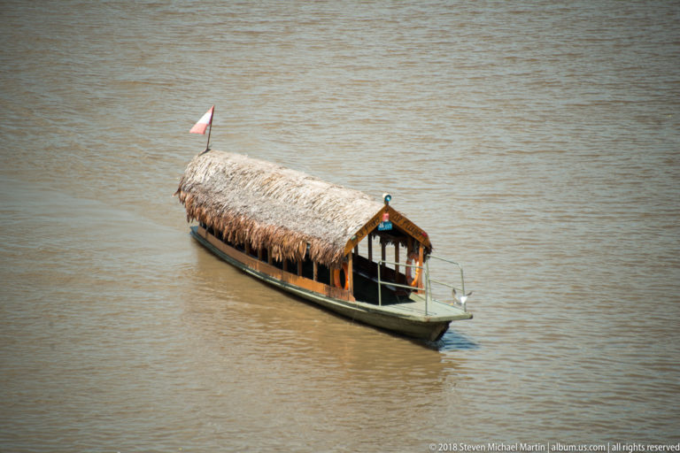 Narrow motorboat with a full palm-thatch canopy and small Peruvian flag traveling across brown river water.