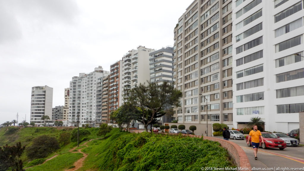 Modern apartment towers lining a green coastal bluff, with a pedestrian in orange walking along the paved path.