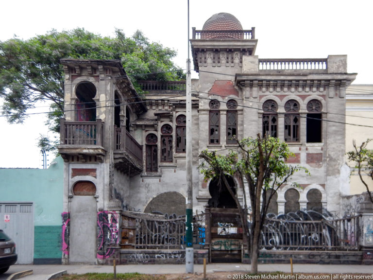 Weathered ornate historic house with broken windows, arched openings, ironwork, graffiti, vegetation, and a small roof dome.