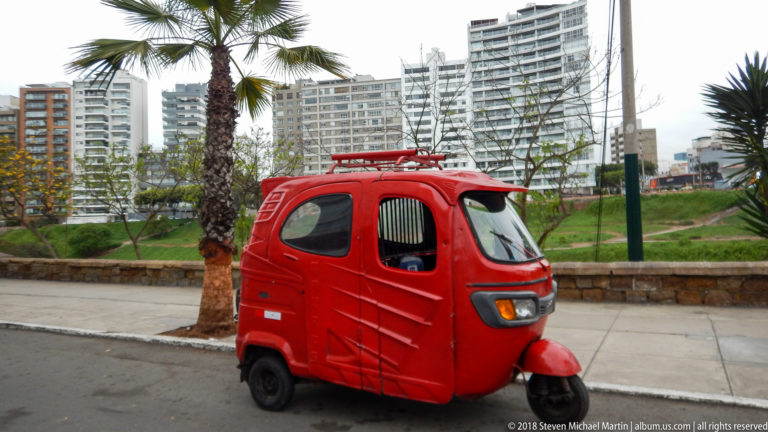 Red enclosed three-wheeled motor taxi parked by a palm, with modern apartment towers across green space behind.