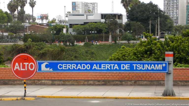 Red ALTO sign beside blue barrier reading CERRADO ALERTA DE TSUNAMI, with greenery and buildings behind.