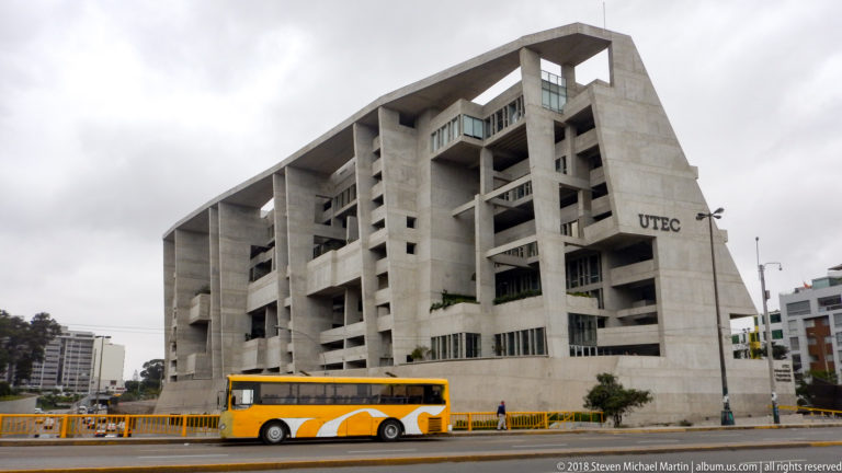 Large exposed-concrete UTEC building with sloping frame, deep terraces, windows, and a yellow bus at street level.