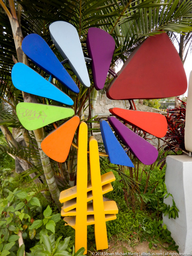 Colorful fan-like abstract sculpture with blue, purple, red, orange, green, and white elements among palm foliage.
