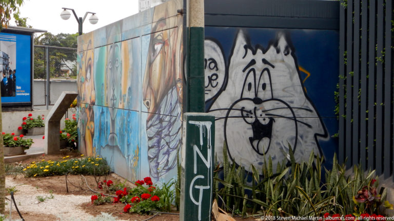 Black-and-white cartoon cat mural on dark blue wall beside multicolored painting, red flowers, and tall green leaves.