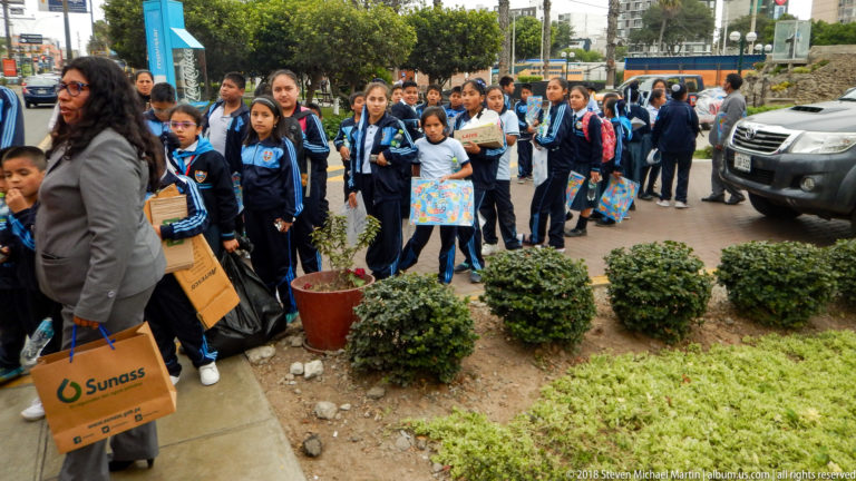 Large group of uniformed schoolchildren with backpacks and materials gathered on a sidewalk beside shrubs and vehicles.