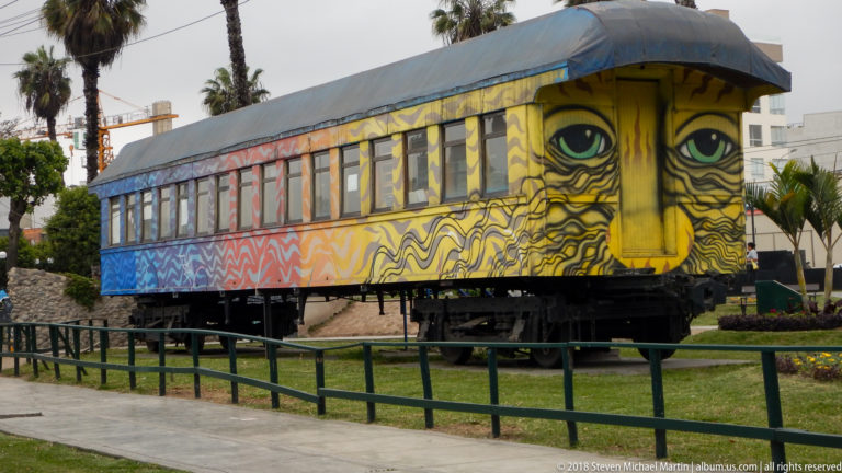 Parked railway carriage covered in colorful mural painting with oversized eyes, displayed behind a low green barrier.