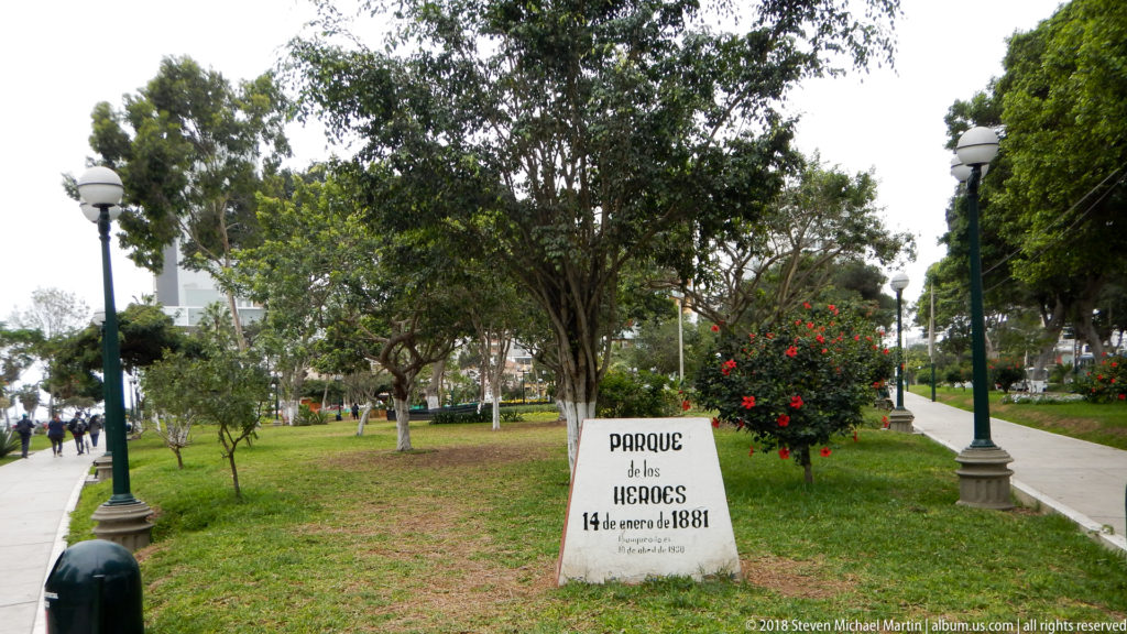 Parque de los Héroes marker dated 14 January 1881 amid lawn, trees, flowering shrubs, lamps, and paved paths.