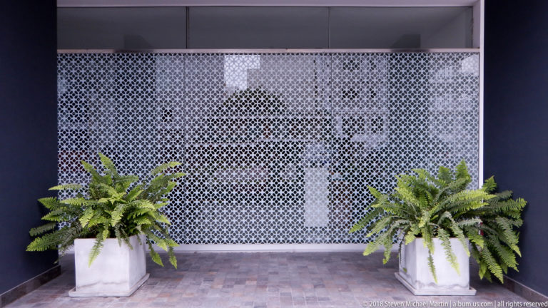 Perforated white architectural screen between dark walls, flanked symmetrically by large ferns in white square planters.