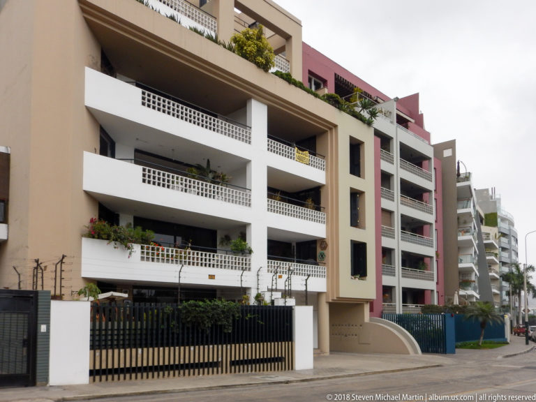 Modern beige, white, and muted red apartment building with deep balconies, plants, and black street fence.