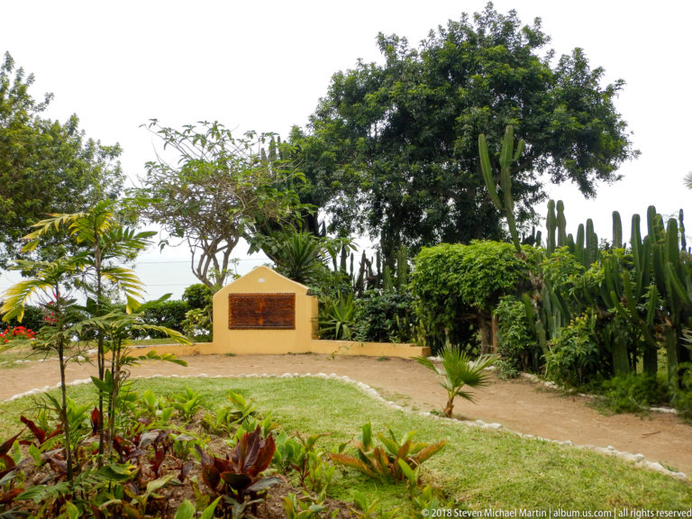 Small ochre memorial marker with dark plaque beside a curving path, shrubs, flowers, and tall cacti.