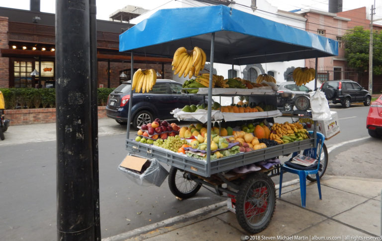 Three-wheeled street fruit cart under blue canopy with hanging bananas and tiers of colorful produce.