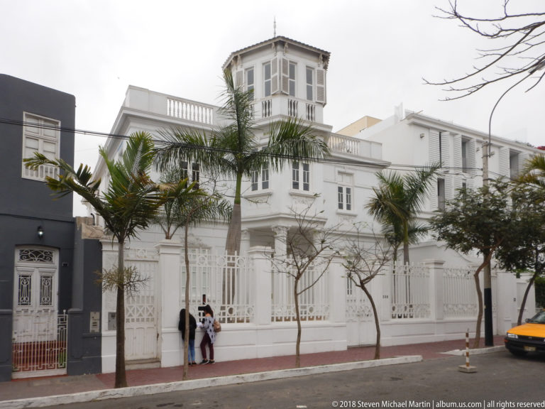 Large white residence behind a metal fence, with tall windows, roof balustrade, palms, and enclosed rooftop lookout.