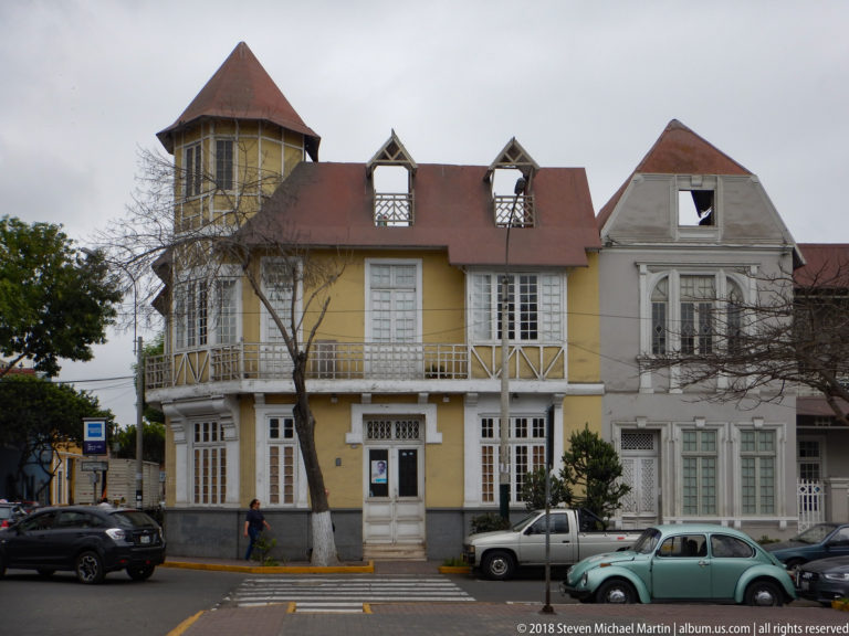 Attached yellow and pale historic residences with red roofs, projecting tower, gables, street trees, and parked cars.