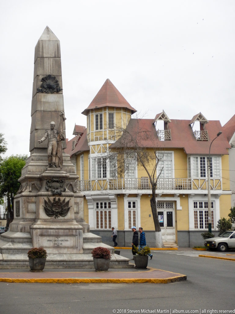 Stone monument with sculpted figures on a traffic island before a yellow building with red roofs and white trim.