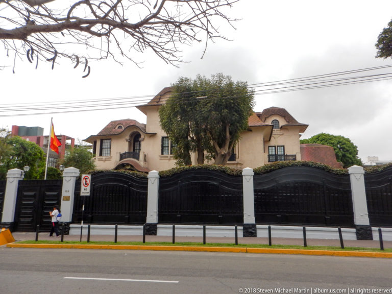 Large cream residence behind tall black gates and white piers, with red tile roofs, balconies, trees, and Peruvian flag.