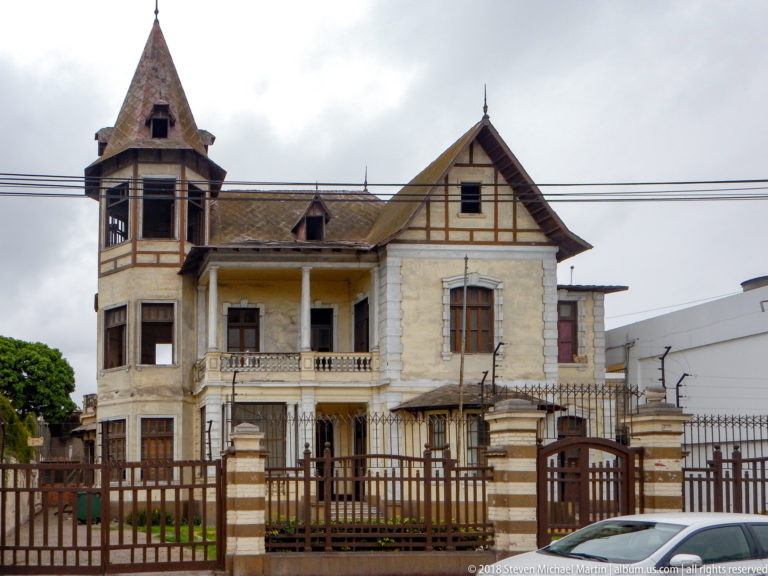 Weathered historic house with square tower, steep gable, timber-like trim, upper balcony, and iron perimeter fence.