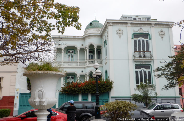 White and mint-green ornate residence with open balconies, decorative windows, small green dome, and large garden urn.