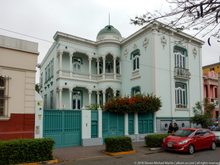Pale blue-green two-story corner house with white trim, columned balcony, tall windows, and matching gate.