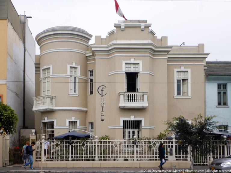 Cream hotel with rounded corner bay, central balcony, white trim, Peruvian flag, umbrellas, and low fence.