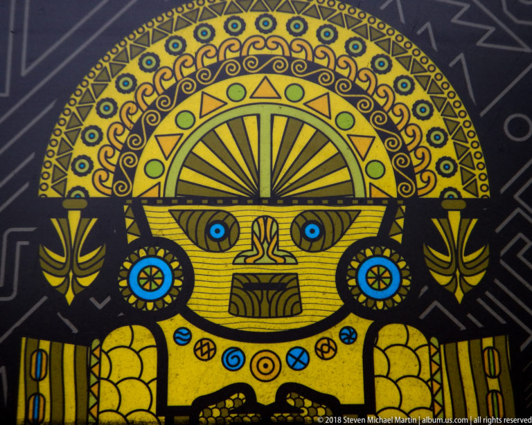 Stylized gold frontal figure with large radiating headdress, blue and green details, and symmetrical motifs on black background.